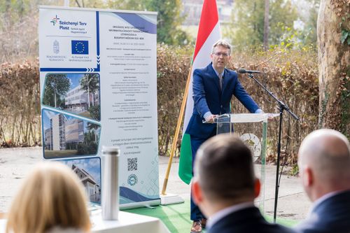 előadó pulpitus előtt, háttérben roll-up és magyar zászló